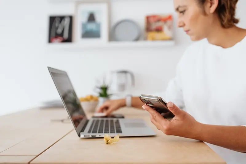 Woman working on a laptop while holding a smartphone at a wooden desk
