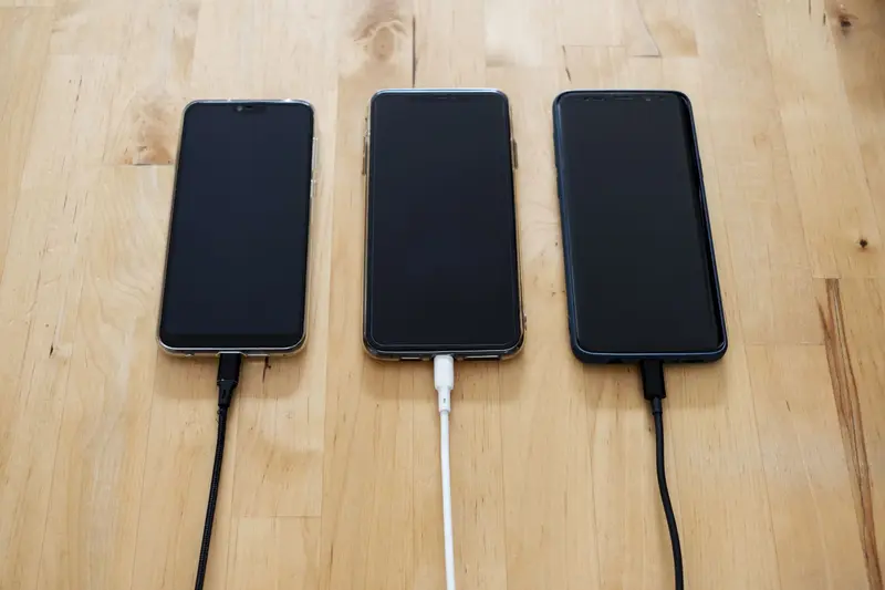 Three smartphones side by side on a wooden table, each plugged into a charging cable